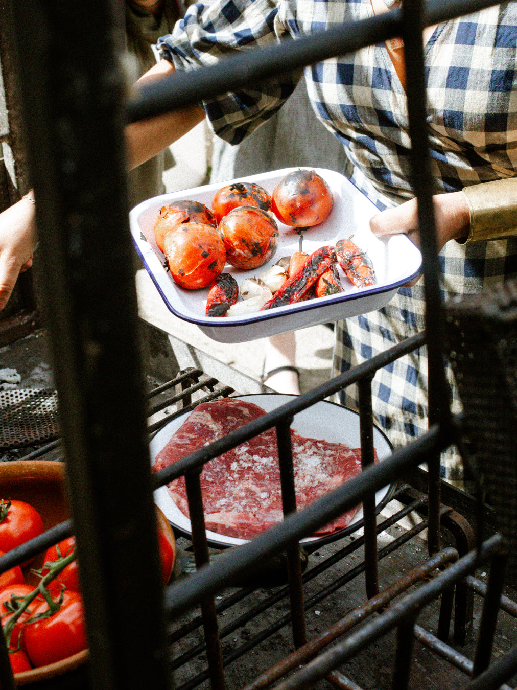 Person holding a tray with grilled tomatoes and raw meat on a grill.