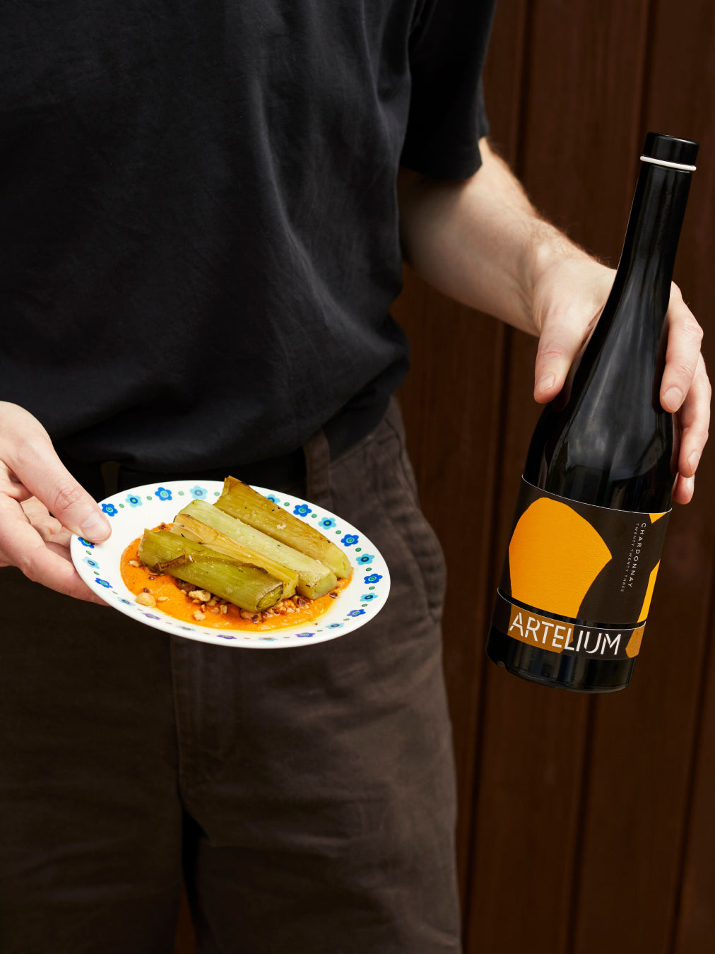 A man holding a bottle of Artelium wine and a small plate with grilled leaks  on a romesco sauce on a brown wood background
