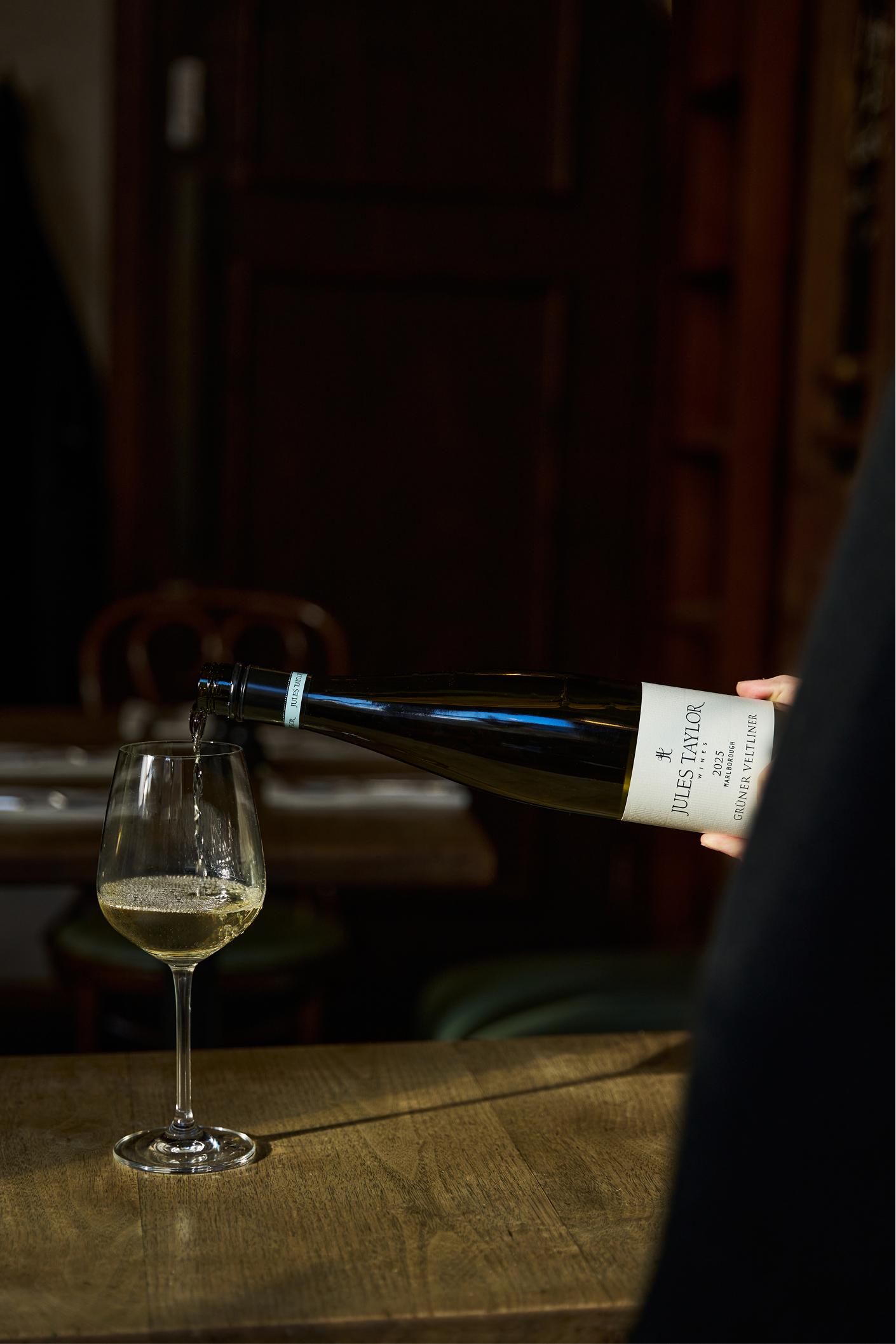 Person pouring Jules Taylor Gruner Veltliner from a bottle into a glass on a wooden table