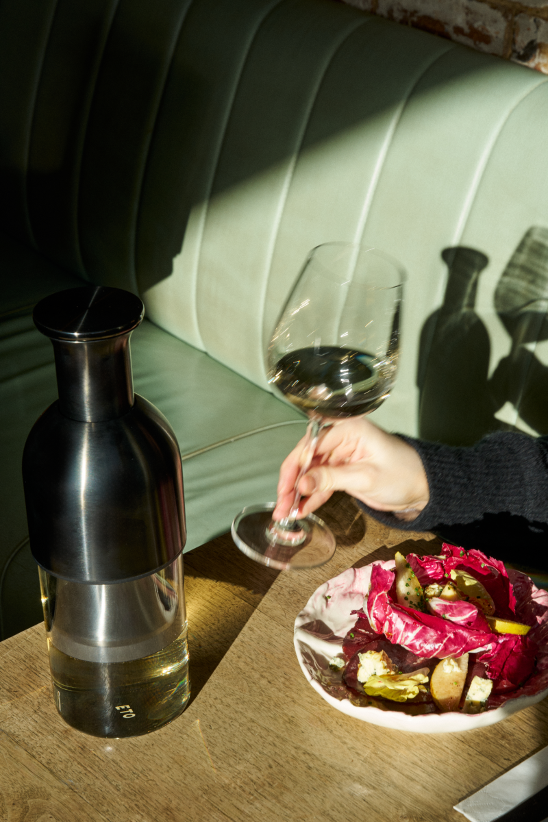 Person holding a wine glass next to a ggraphite decanter and a plate of food on a table.