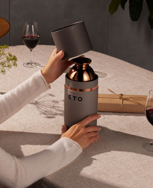 Person holding a wine bottle with a gray and copper design, labeled 'ETO', on a table with glasses and a gift box.